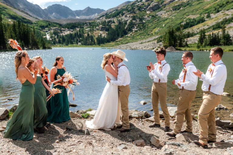 Couple kissing by Colorado Alpine lake with their wedding party cheering them on.