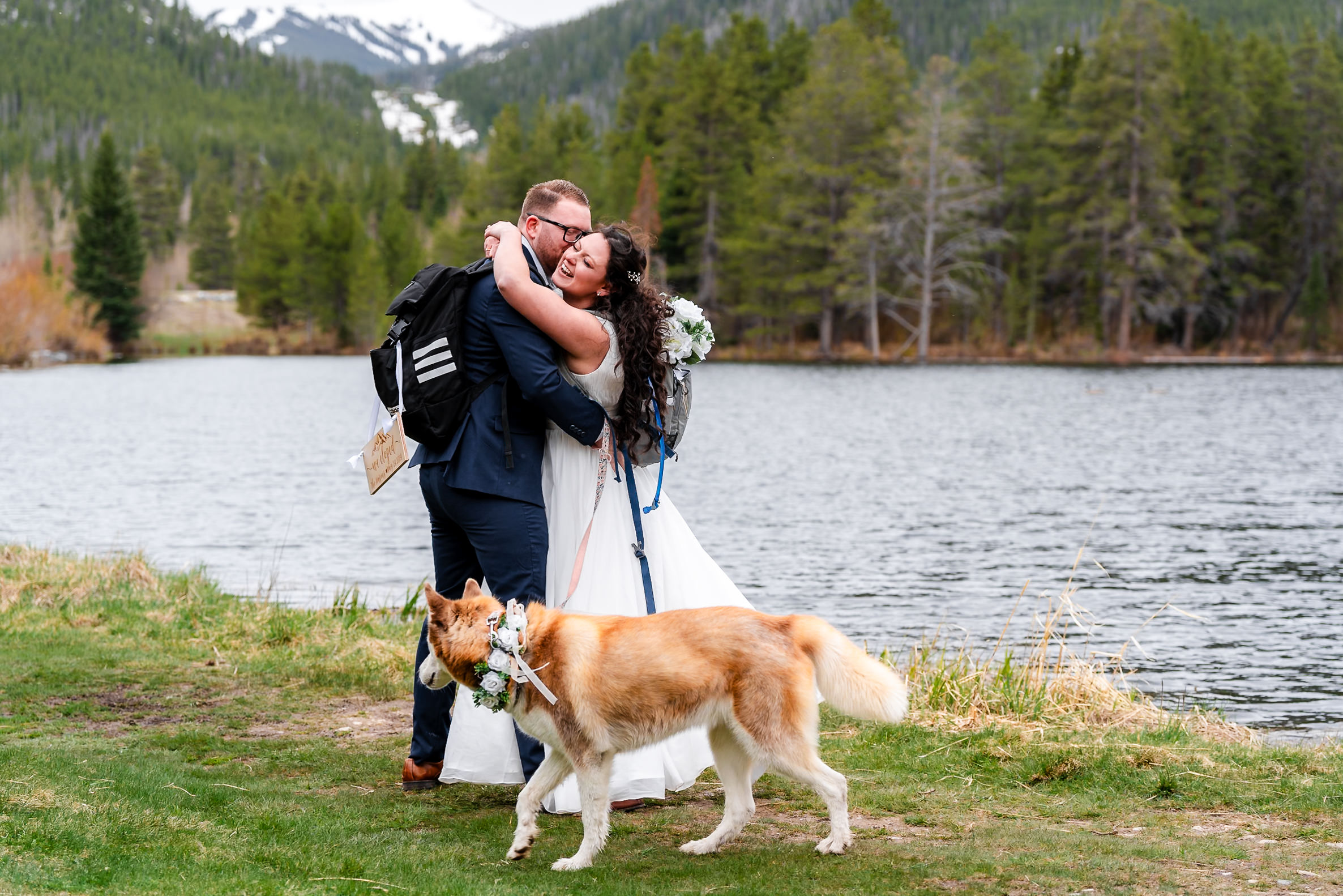 groom and bride hugging by a lake with their dog standing in front of them