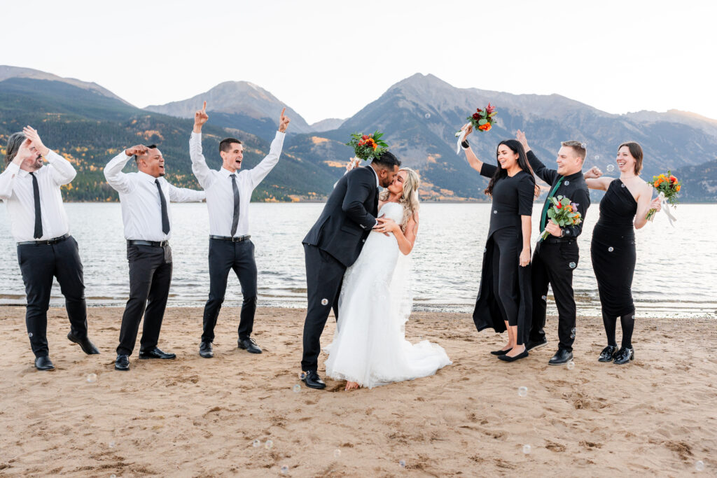 a bride and groom kissing surrounded by a cheering wedding party on a beach in front of a lake and scenic mountains