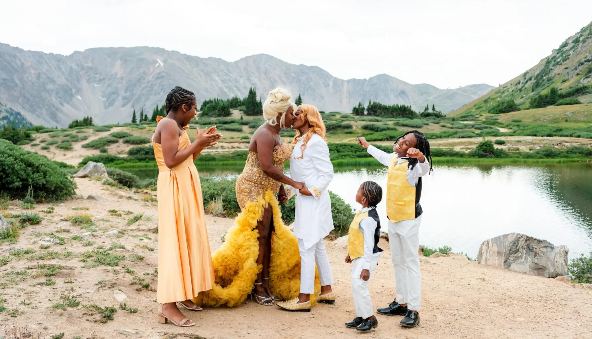 Couple sharing a kiss by an alpine lake as their family celebrates during a Colorado mountain elopement