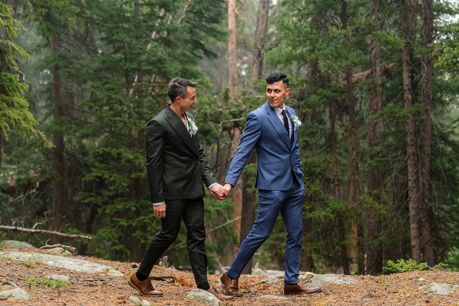 Two grooms walk hand in hand through a Colorado mountain forest during their outdoor elopement ceremony