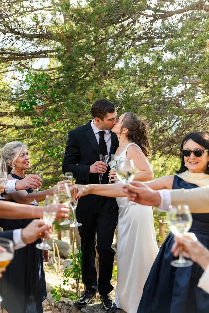 Bride and groom kissing as guests raise glasses in a champagne toast at an intimate outdoor Colorado wedding