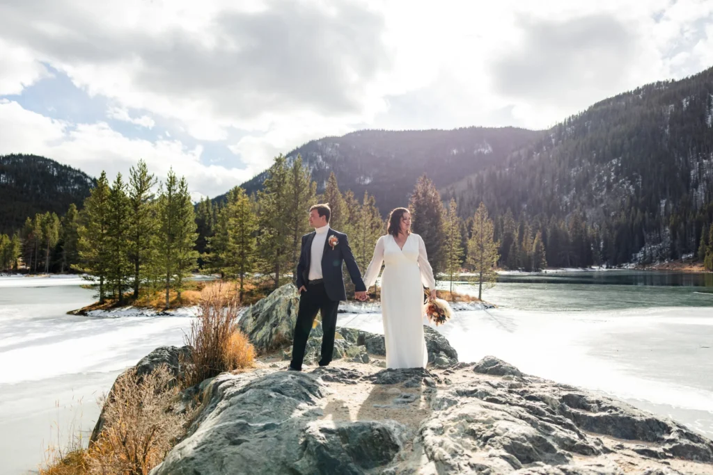 Bride and groom holding hands on a rocky outcrop over a frozen mountain lake at their winter Colorado elopement